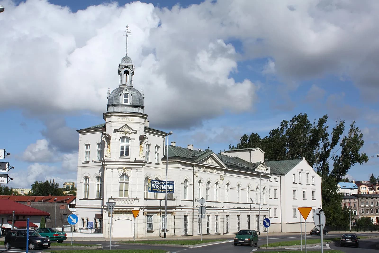 A grand white building with a domed tower on the left, featuring multiple windows and architectural details, stands prominently next to a road with a few cars and trees in the background under a partly cloudy sky.
