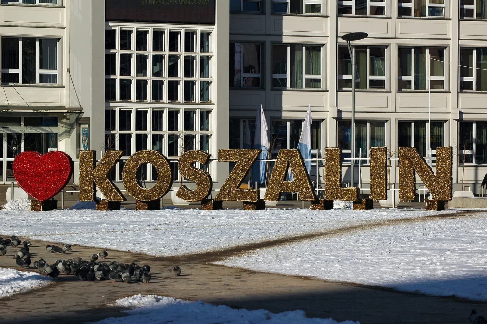 A large red heart and glittery gold letters spelling KOSZALIN stand in a snowy public square.